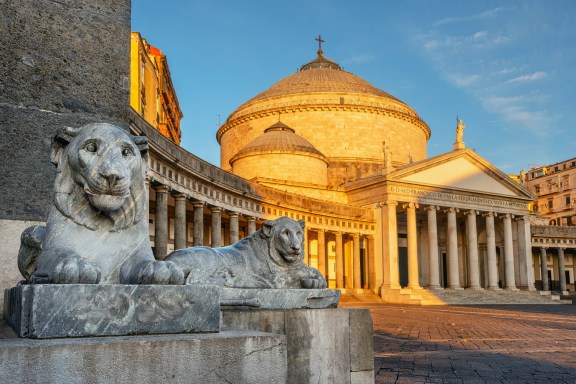 Two lion statues in the foreground and behind it a beautiful old church bathed in warm sunlight in Naples